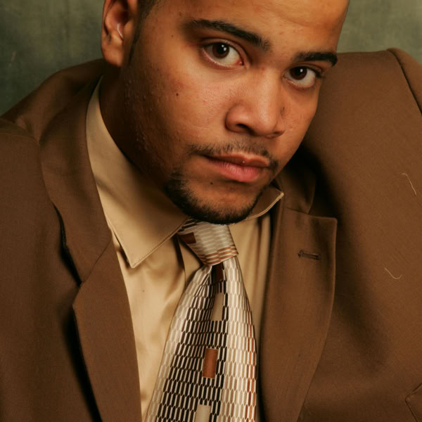 Headshot of a man with stubble in a brown suit and tie. Indoor setting.